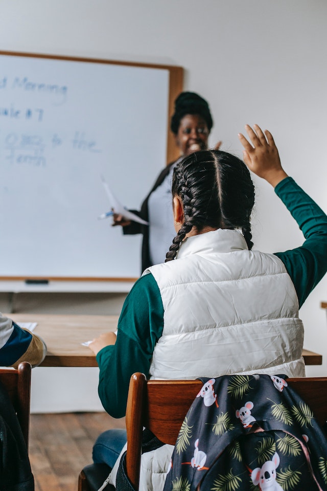 Classroom setting with teacher in front of whiteboard and student with raised hand. By Katerina Holmes on Pexels